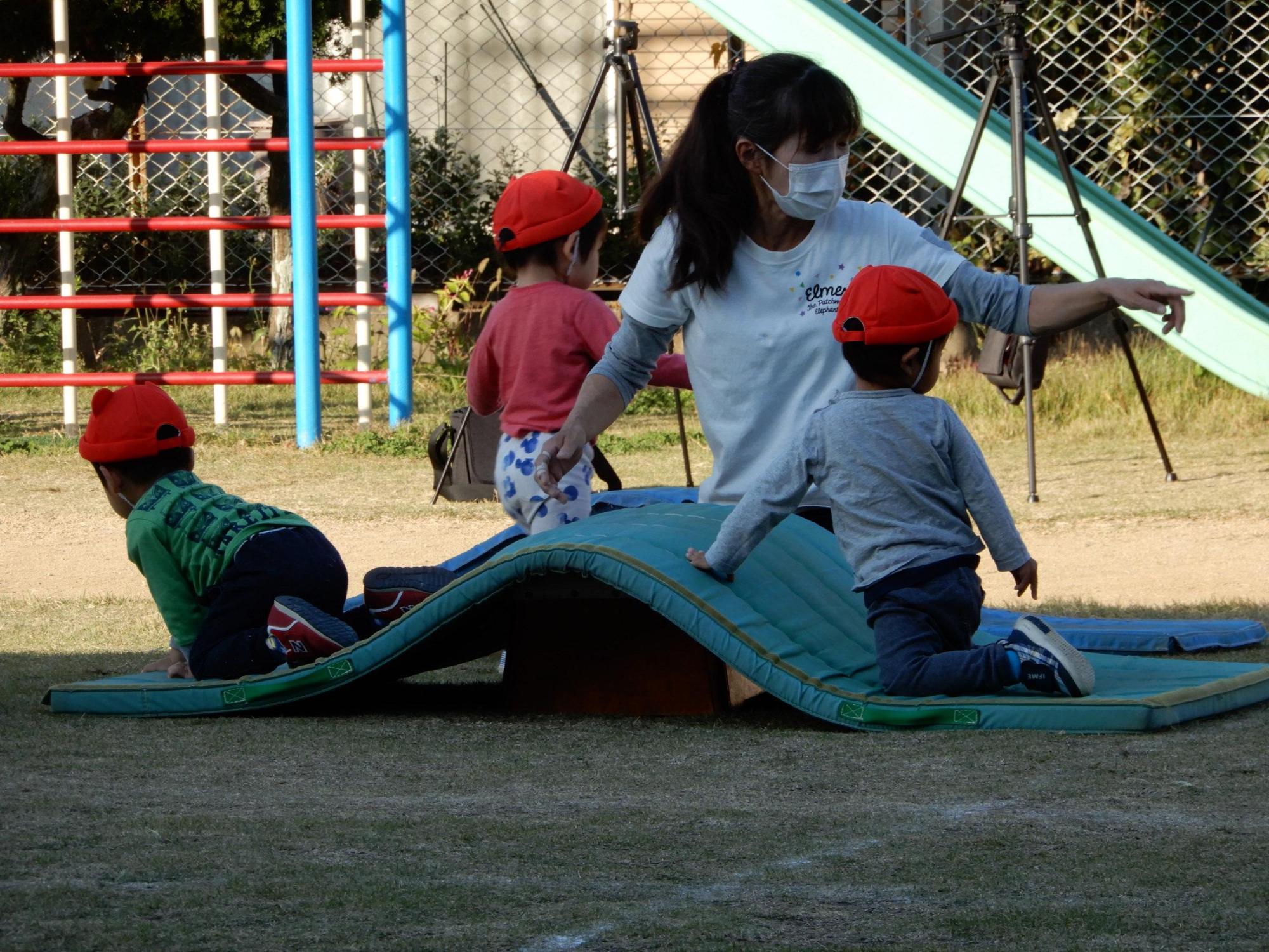 障害物競走で1段の跳び箱の上に置かれたマットを降りる男児の後ろで先生が左手人差し指で指す方を向く男児の写真