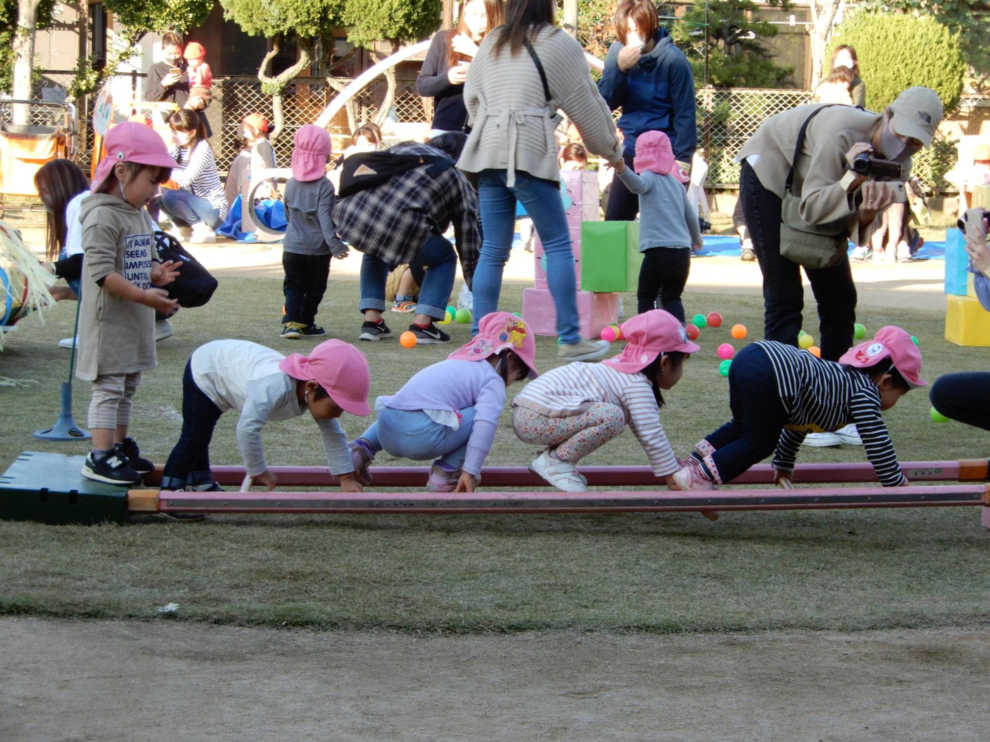 障害物競走で横に倒したはしごの段を掴みながら前進する園児4人の様子をカメラで撮影する保護者の写真