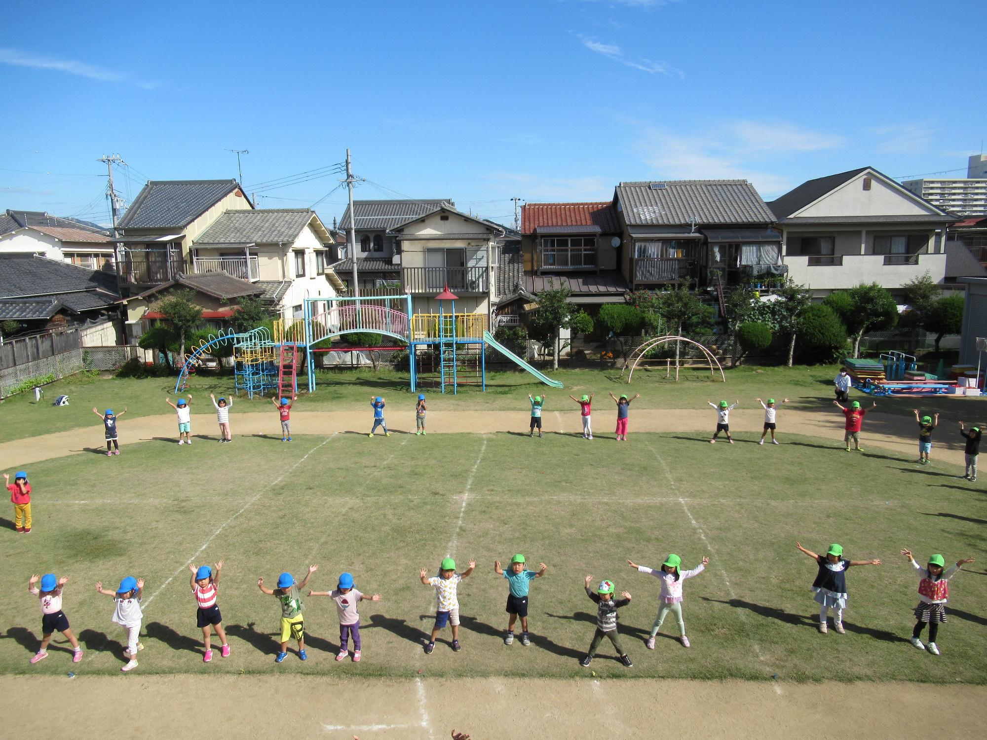 園庭で輪になり全身を使って体操をする園児たちを上空から撮影した写真
