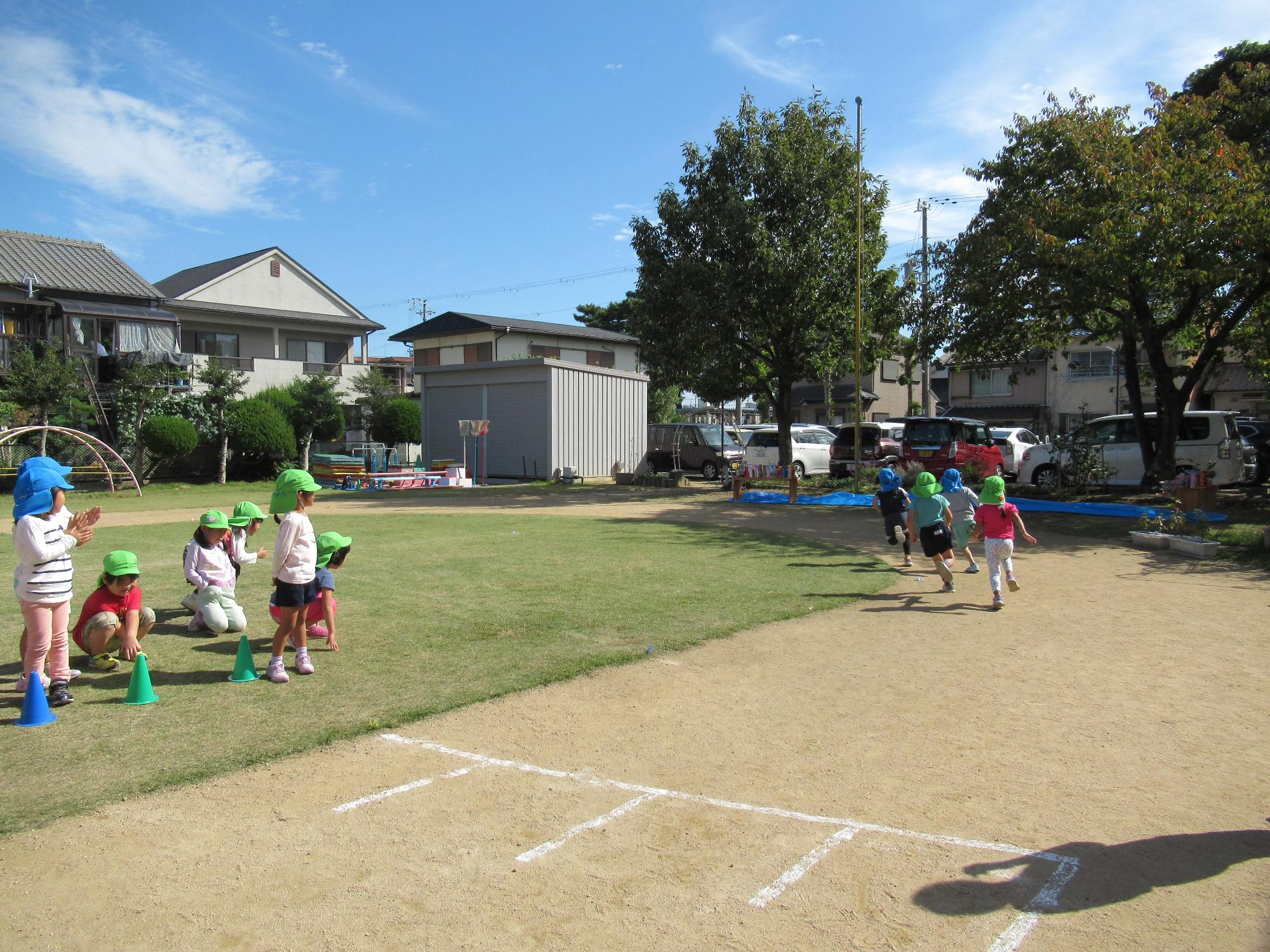 白線で引かれたゴールを目指し走る園児たちとゴールの右横で手を叩いて応援する園児たちの写真