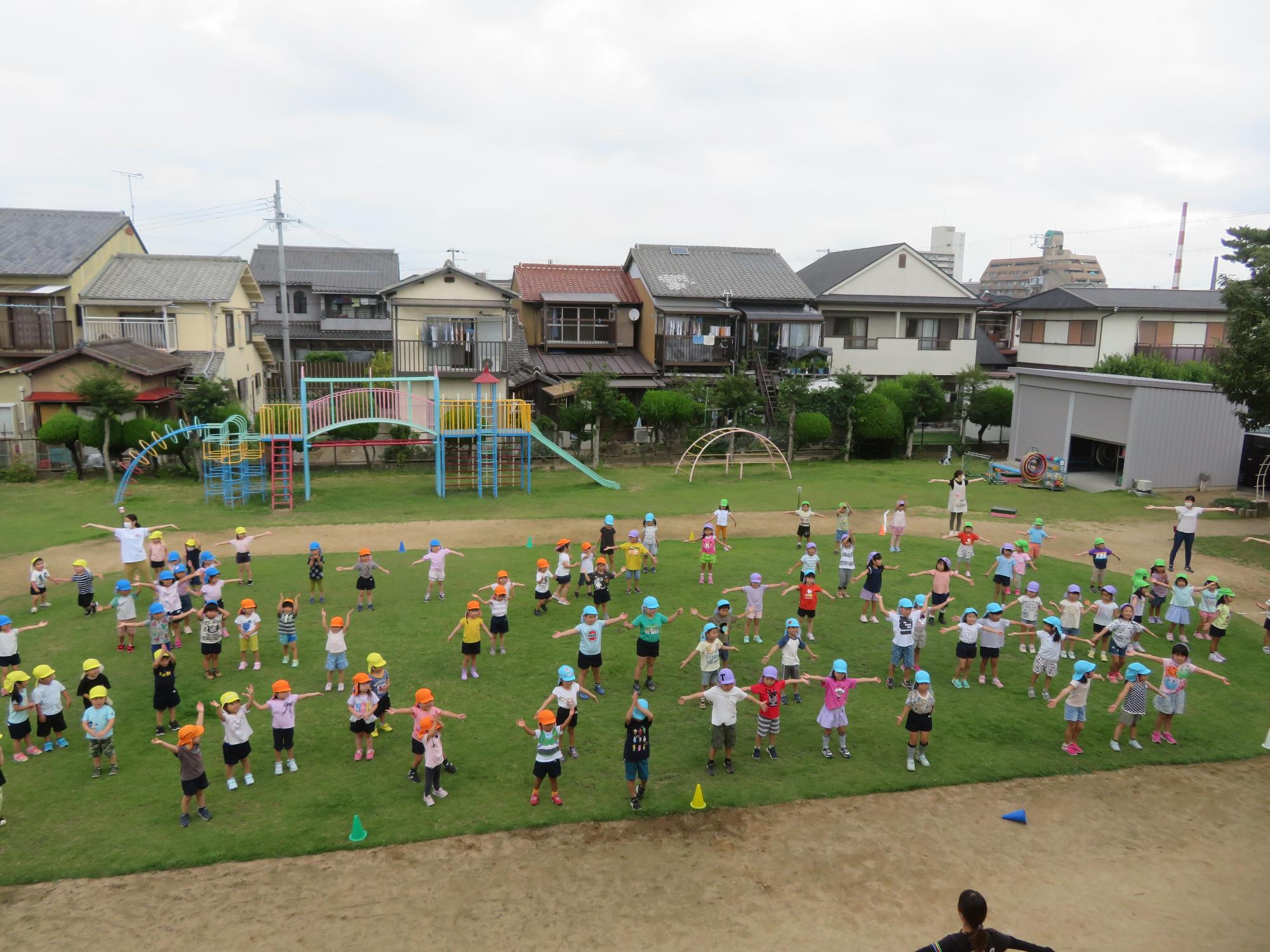 園庭で先生たちと一緒に両手を広げて大好きニッポンを体操する園児たちの写真