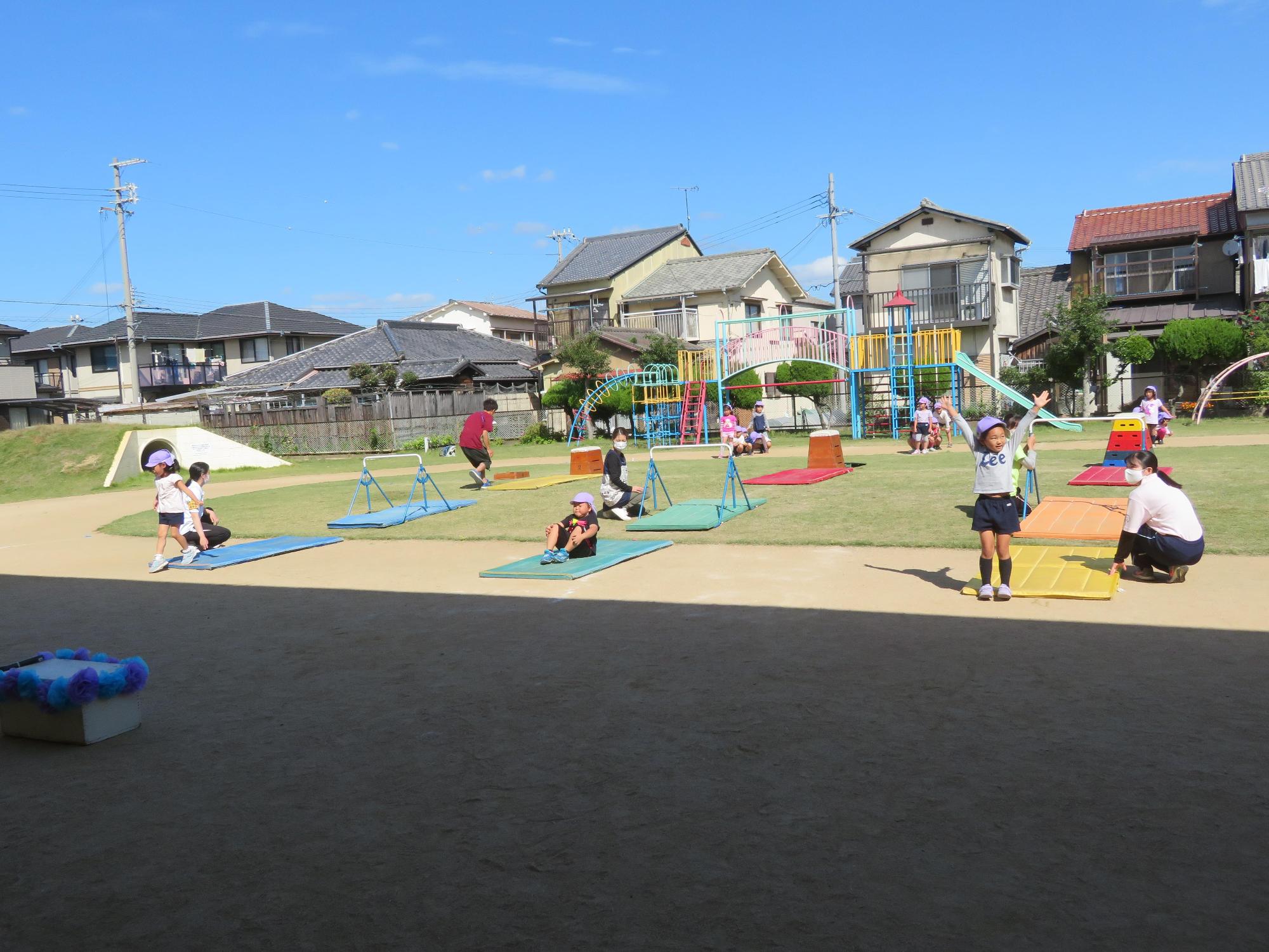 先生に手伝ってもらいながら3段の跳び箱鉄棒マットの順で遊ぶ園児たちの写真