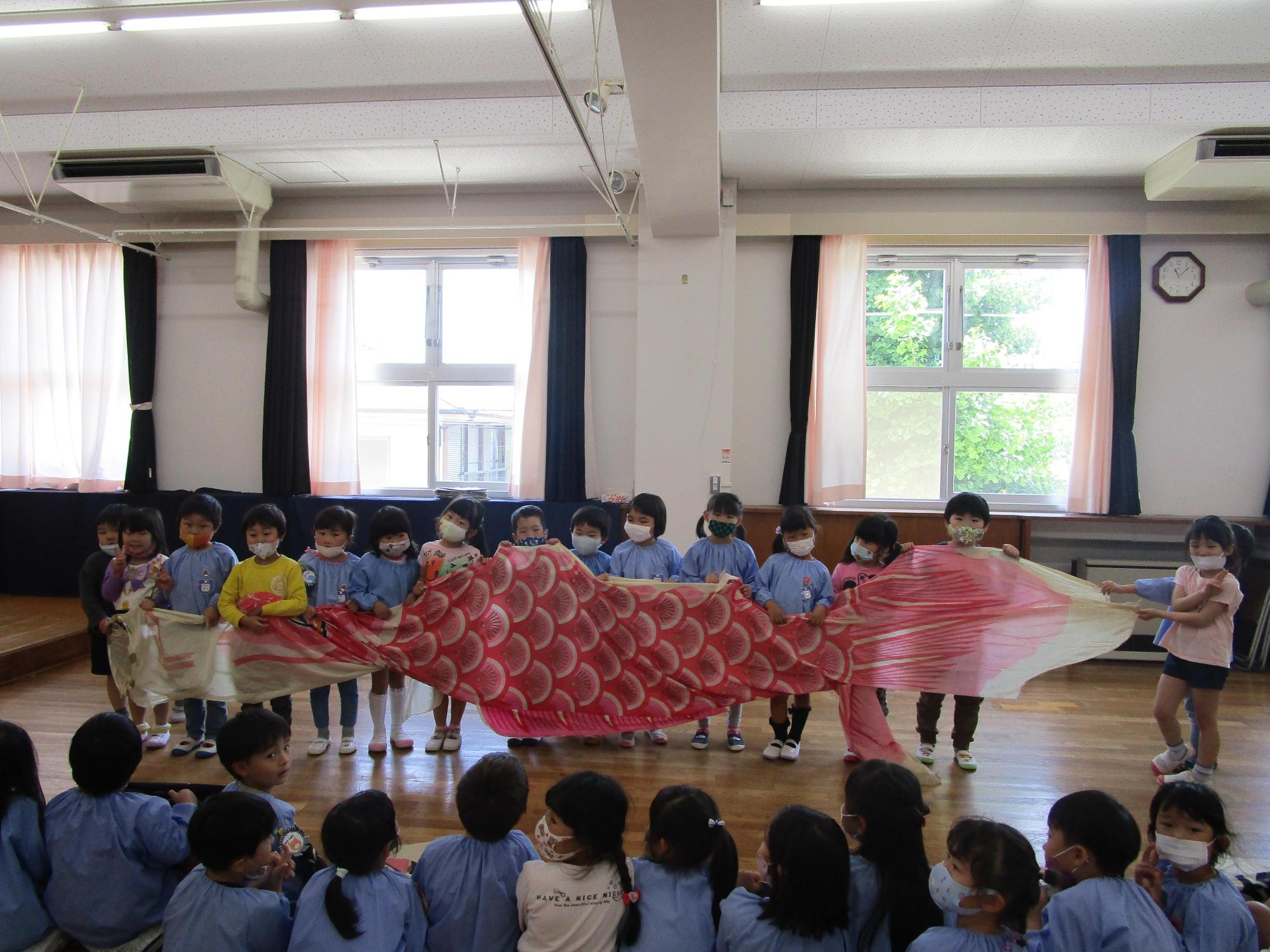 フローリングの大きな部屋で約10名ほどの保育園児たちが横一列に並び大きな赤いこいのぼりをみんなで広げて持っておりそれを他の保育園児たちが向かいに座って眺めている様子の写真