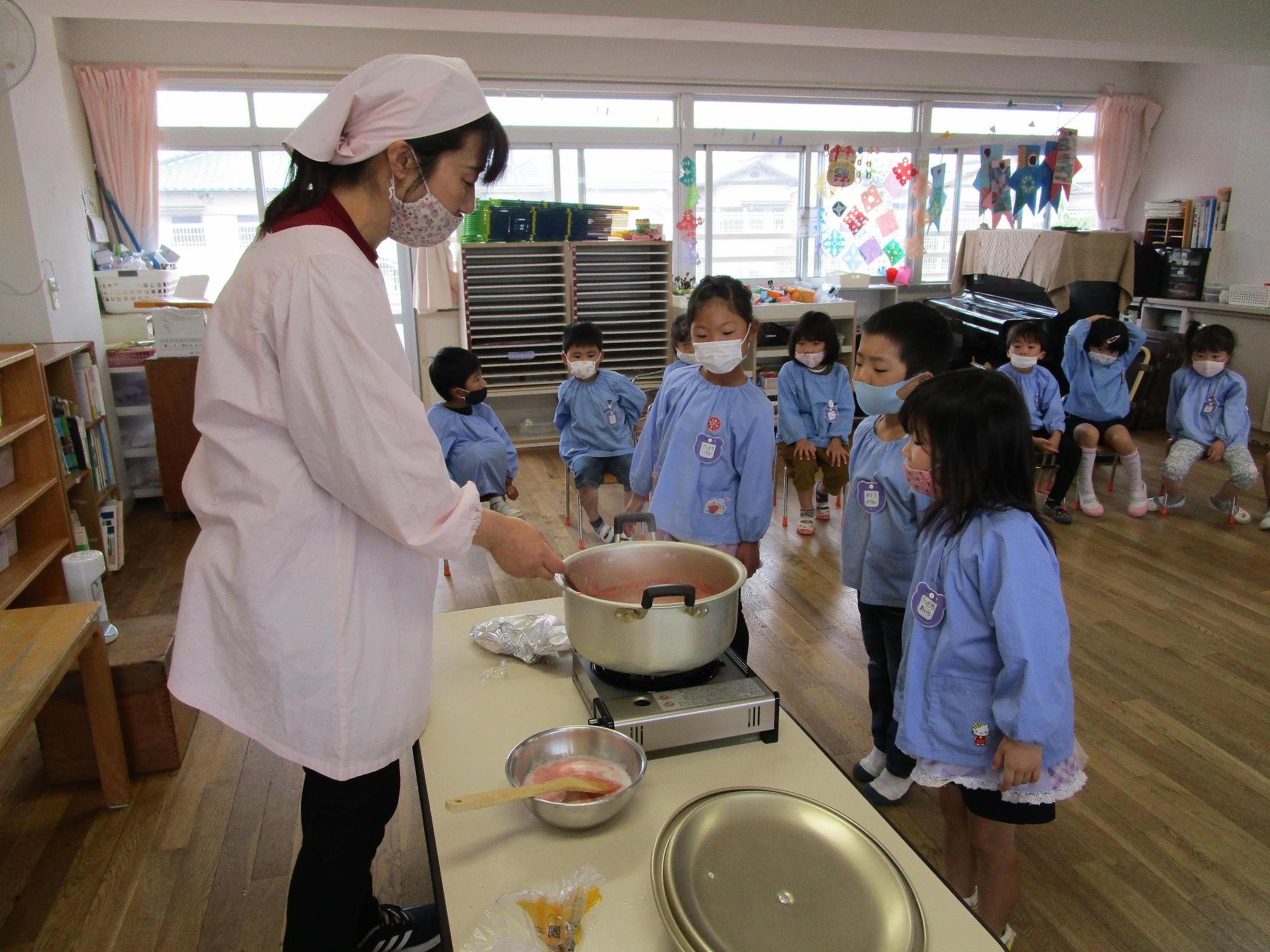 保育園のスタッフがイチゴと砂糖とレモン汁を鍋で煮詰めているところに近づき鍋を覗き込もうとしている3人の園児と少し離れた場所に座っている数名の園児の写真