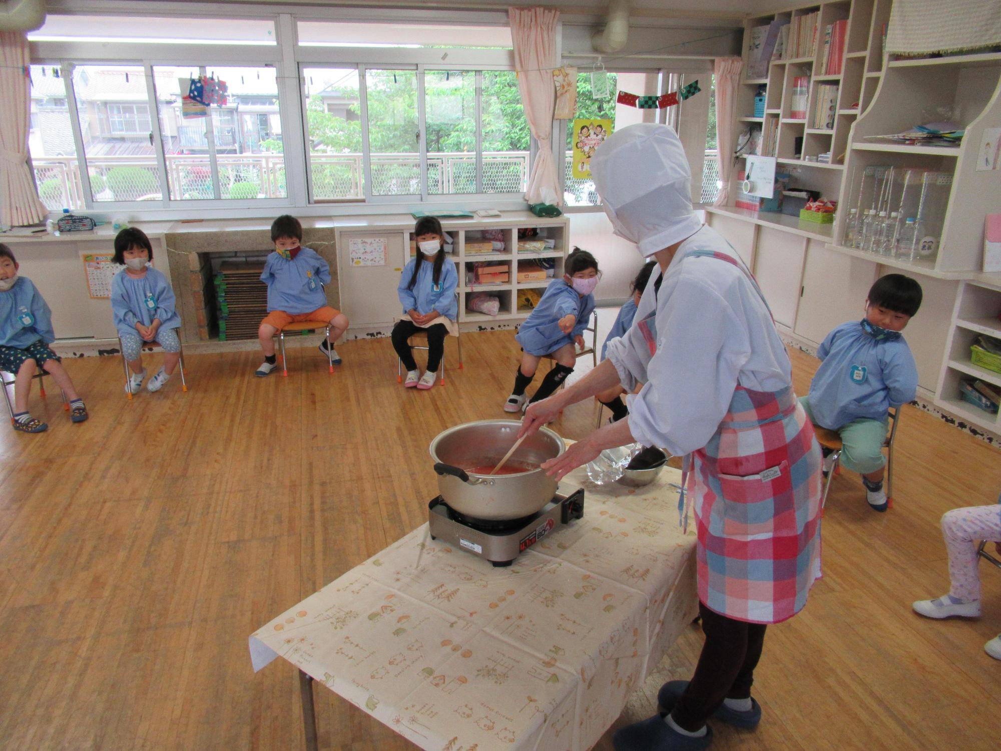 保育園のスタッフがイチゴと砂糖とレモン汁を鍋で煮詰めているのを保育園児たちが少し離れた場所から椅子に座って見守っている様子の写真