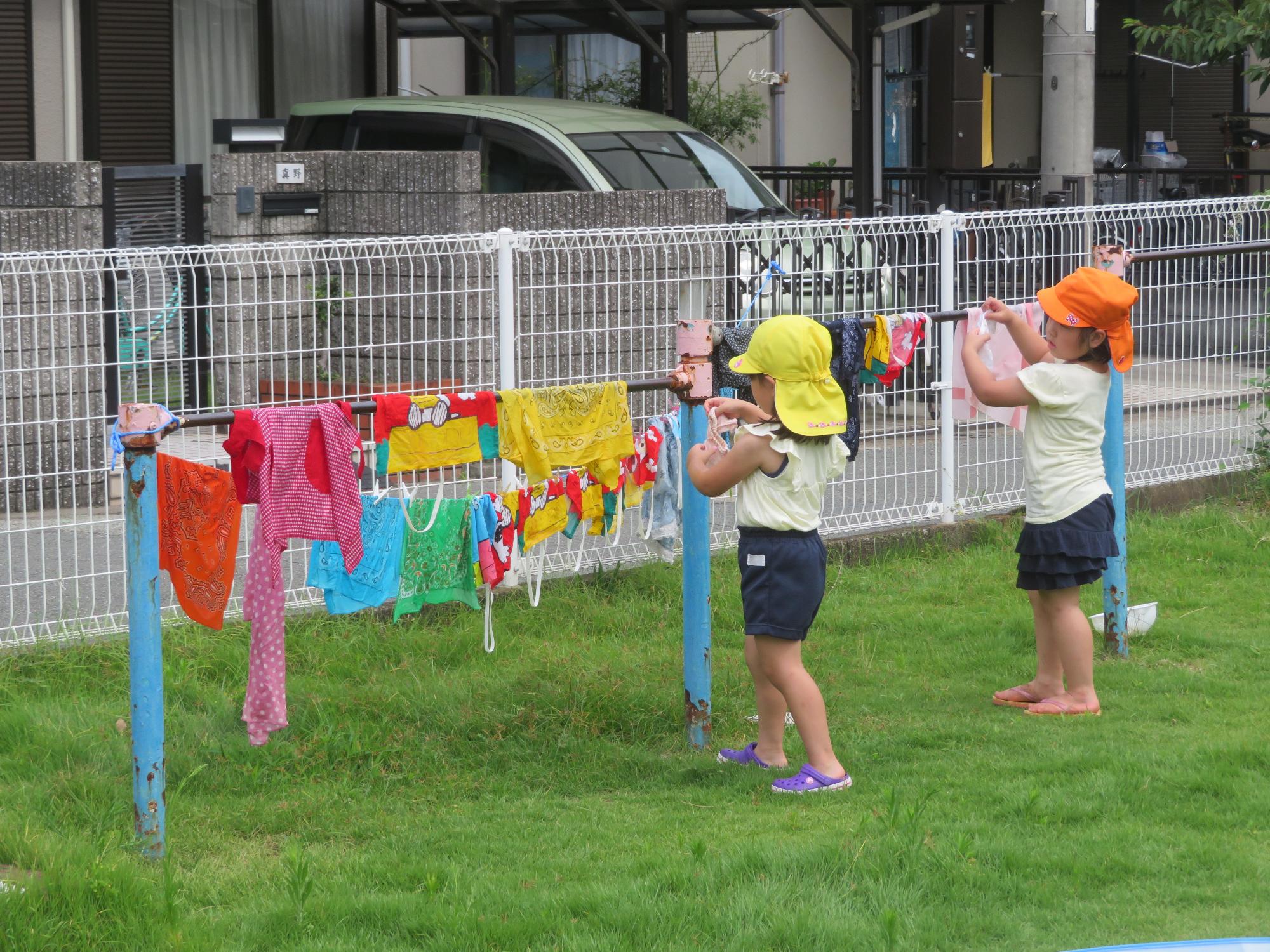 園庭にある鉄棒に靴下を干そうとしている女児の右横で洗濯物を干しながら心配そうにその様子を見守る女児の写真