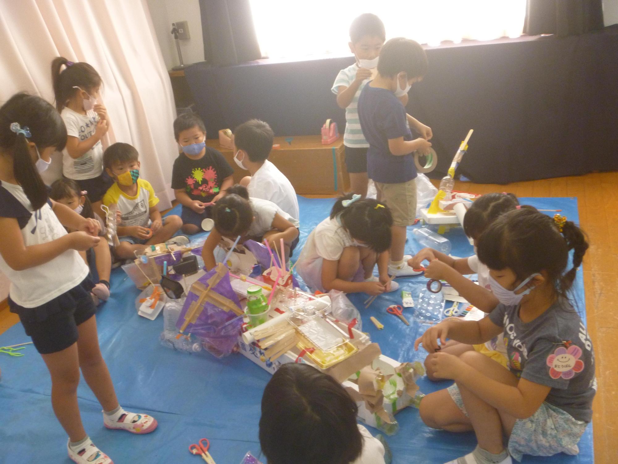 ブルーシートの上で廃材を使ってみんなで協力しながら船作りをする5歳児クラスの園児たちの写真