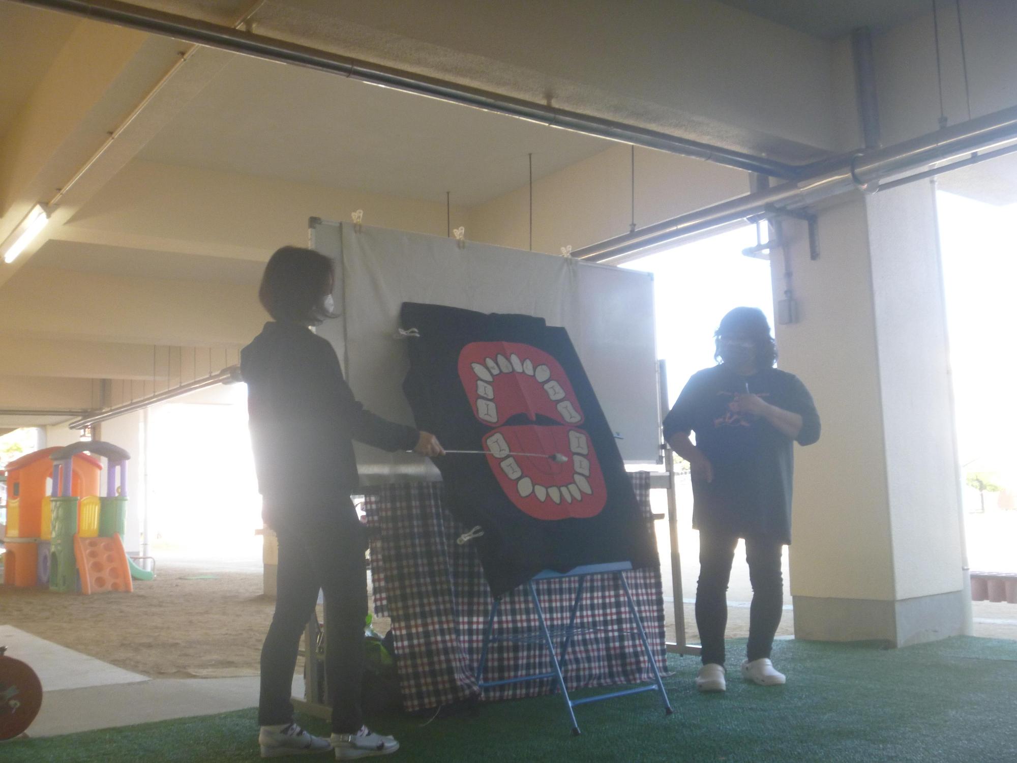 建物のピロティ部分の少し暗がりになっている場所で2人のスタッフが歯のイラストを指示棒で示しながら歯科健康教育を行っている様子の写真