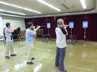 地区公民館内の一室で等間隔に並べられた的に向かって矢を吹く男性3人を背後から撮影した写真