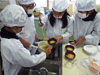 すでに麺がよそってあるお椀に鍋からスープを入れようとする児童たちの写真。全員が割烹着を着ている。
