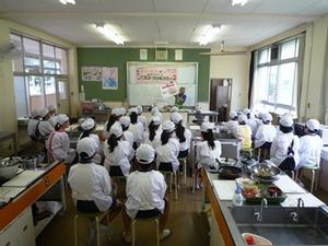 米田西小学校の家庭科室にて、ホワイトボードの前で説明する講師と、それぞれスツールに座って話を聞く児童たちの写真