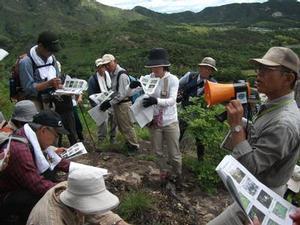 高御位山にて、植物の写真が並んでいる資料を見ながら先生の説明を聞く参加者たちの写真