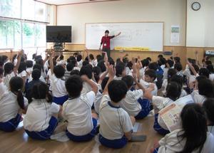 米田小学校内の特別教室で、壁面に備え付けられたホワイトボードを使って説明する職員と、それぞれ床に座って話を聞く児童たちの写真。職員の問いかけに手を挙げて答えている。