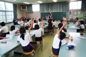 荒井小学校内の特別教室にて、黒板の前で資料を高く掲げる市の職員と、長机の周囲のスツールに座りながら挙手する児童たちの写真
