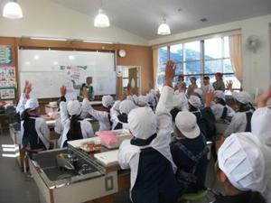 阿弥陀小学校の家庭科室にて、教室壁面のホワイトボードに貼られたパネルの指差す講師に対して、割烹着姿の児童たちが全員手を挙げている。