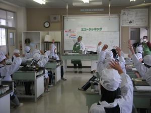 曽根小学校の家庭科室にて、講師が掲げながら見せている大判のパネルに対して、割烹着姿の児童全員が手を挙げている写真