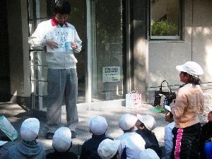 米田小学校の屋外で資料を見せながら話をする講師とそれを地面に座って聞く児童たちの写真。児童の1人が立って講師と話をしている。