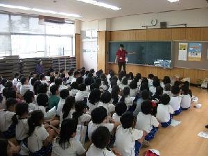 米田小学校の特別教室で黒板を使って話をする市の職員と、それを床に座って聞く児童たちの写真