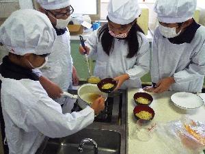 中筋小学校の家庭科室にて、麺を盛り付けたお椀に鍋のスープを入れようとしている児童たちの写真
