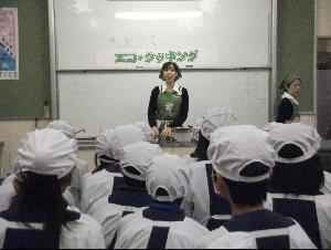 米田西小学校の家庭科室にて、壁面のホワイトボードの前で話をする講師と、それを聞く割烹着姿の児童たちの写真