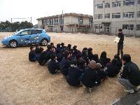 校庭で電気自動車についての話を聞く生徒たちの写真。校庭の奥には電気自動車である公用車の宝くじ号が停まっている。