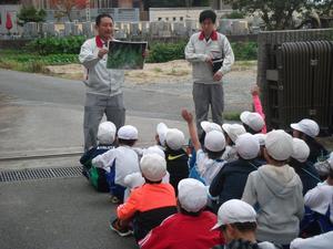米田小学校での自然観察会の写真で、屋外にて写真を見せながら説明する講師と、その話を地面に座りながら聞く児童たちが撮影されている。