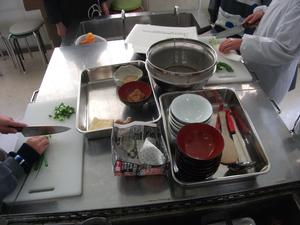 曽根小学校でのエコクッキングにて、これから使う笊やしゃもじといった調理器具が並んでいる写真。その脇では包丁で食材を切り始めている。