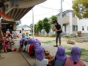 梅井保育園の園庭で資料を見せながら話をする市の職員と、その話を体育座りをしながら聞く園児たちの写真