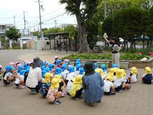 荒井幼稚園の中庭で資料を掲げながら話をする市の職員と、それをしゃがみながら聞く園児たちの写真