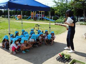 曽根幼稚園の園庭にて立って資料を手にしながら話をする職員と体育座りで話を聞く園児たちの写真。園児たちはイベント用テントの下にいる。