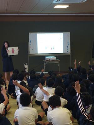 阿弥陀小学校の屋内でホワイトボードをスクリーン代わりにしてプロジェクターを使いながら環境学習について話す講師と、床に座ってその話を聞く児童たちの写真。講師が資料を手にしながらした問いかけに何人かの児童が手を挙げている。