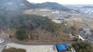 手前に道路があり、真ん中に地肌が少し見える山があり、奥に畑がある写真