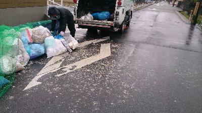 1人のゴミ収集員が道路の端に散乱しているゴミ袋をゴミ収集車に詰め込もうとしているが、そこから漏れた液体物が周囲を汚してしまっている写真