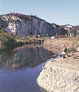 小川にいる釣り人越しに見る龍山の絶壁部の写真