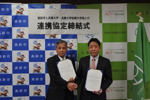 高砂市長と兵庫大学・兵庫大学短期大学部の職員の男性が協定書を持って握手をしている写真