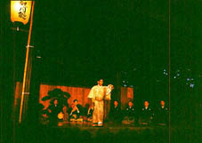 暗い舞台に浮かび上がっている、並んで座った演者達と前に立つ和服の男性の写真