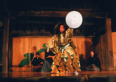 銀色の丸い月を模した板を手に掲げている、和服の装束を着て面とかつらをつけた男性の写真