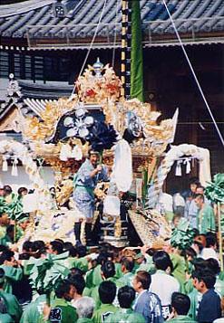 黒地に金色の装飾がほどこされた華やかな神輿に乗る人と、周りに担いでいる人々の活気ある写真