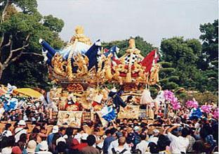 奥には赤い飾りの神輿、手前には青い神輿が大勢の人々に担がれている写真