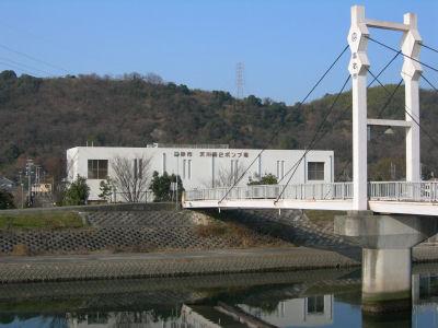 橋の架かった先に建つ白い建物の天川第2ポンプ場の外観写真