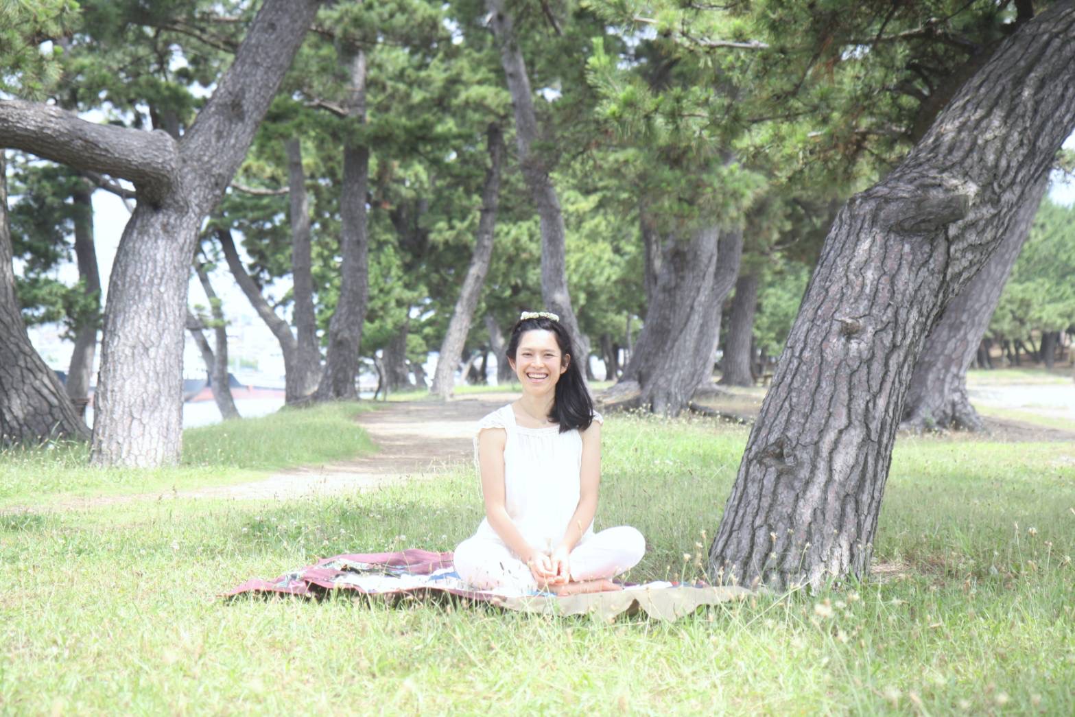 松林の中で、芝生の上に敷物を敷いて座る白い服の女性