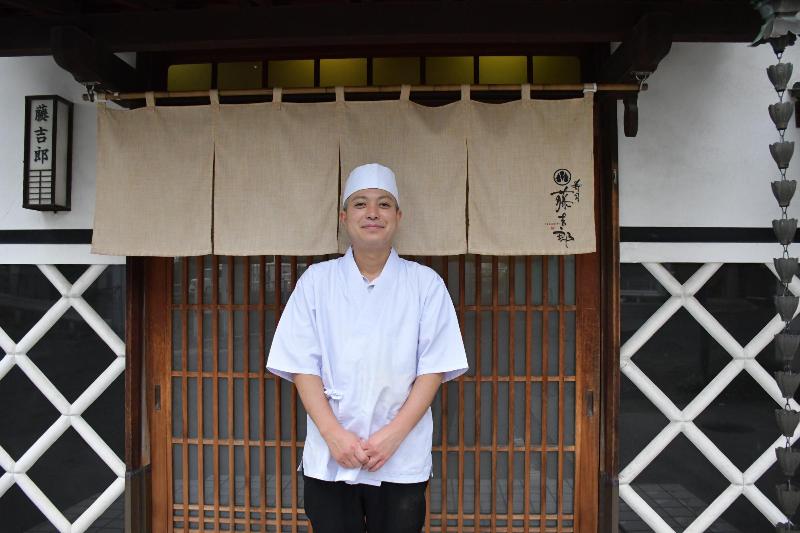 寿司屋ののれんの前に立つ白い服の男性の写真