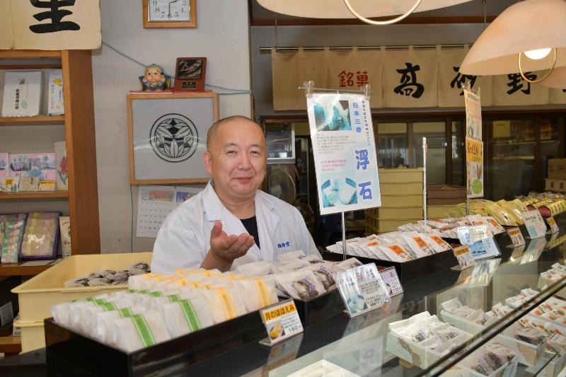 和菓子が陳列されたカウンターの中で微笑みながら手を差し出す白い服の男性