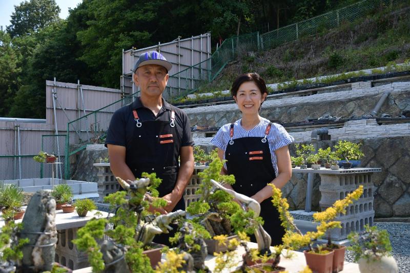 たくさんの盆栽に囲まれて立つ男女の写真