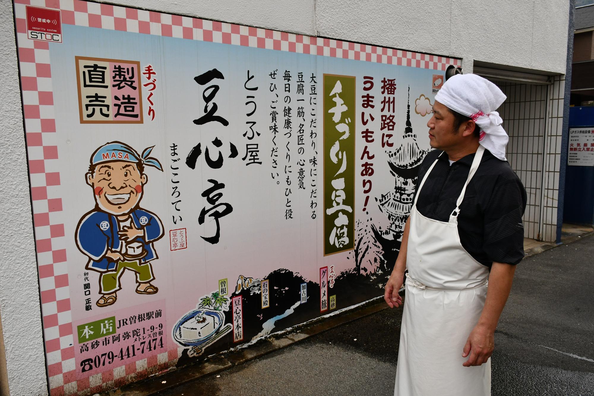 先代が描かれた看板の前に立つ永田さん