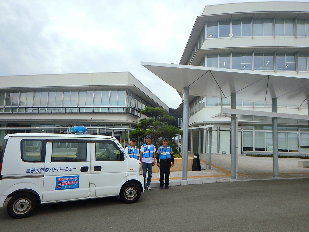 高砂市役所の前に止まった青色防犯パトロール車の前にそれぞれ青色の上着を着た3人の青色防犯パトロール員が並んでいる様子の写真