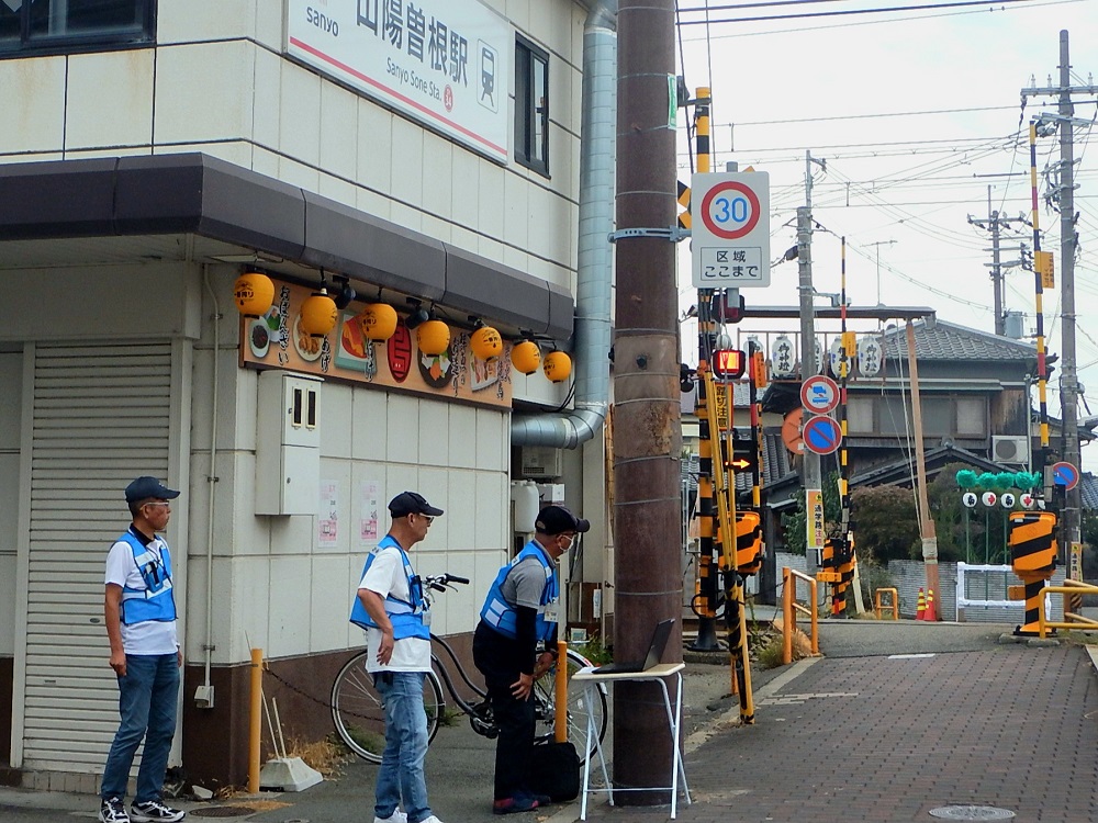 踏切の近くに設置されている見守りカメラの設備点検を行う3名の青色防犯パトロール員のようす